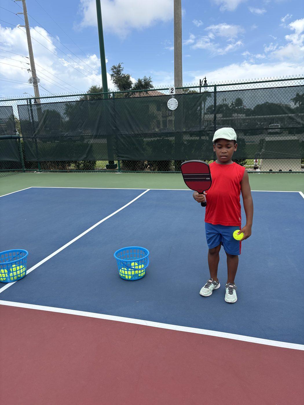 A young player holding a paddle and ball beside baskets of pickleballs.