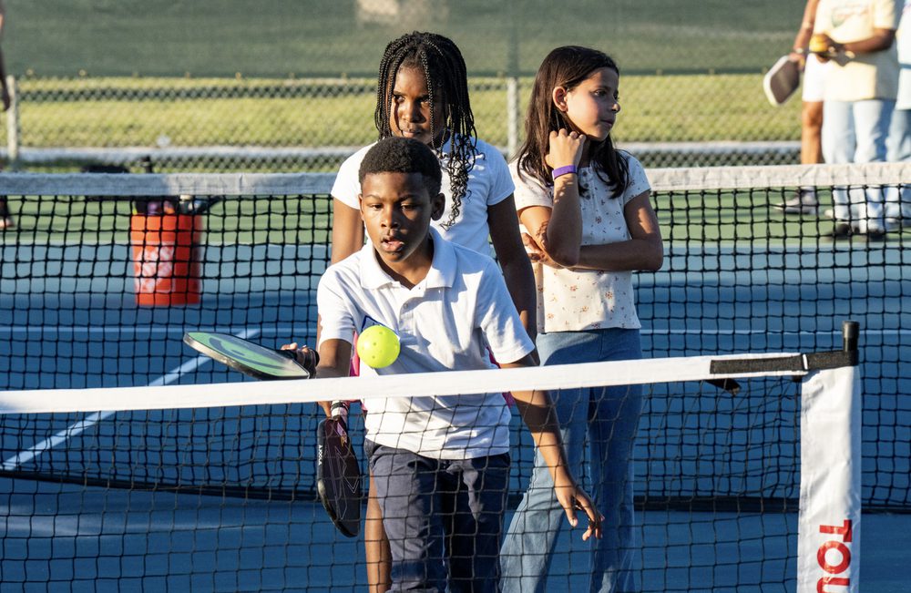 Three young players standing at the net, one holding a ball and paddle.