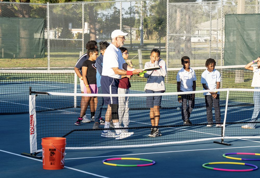 A coach demonstrating to a line of kids at the net, hula hoops marking the station.
