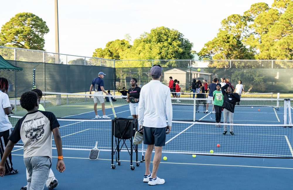 A boy walking onto a blue court while a coach sets up a ball cart.