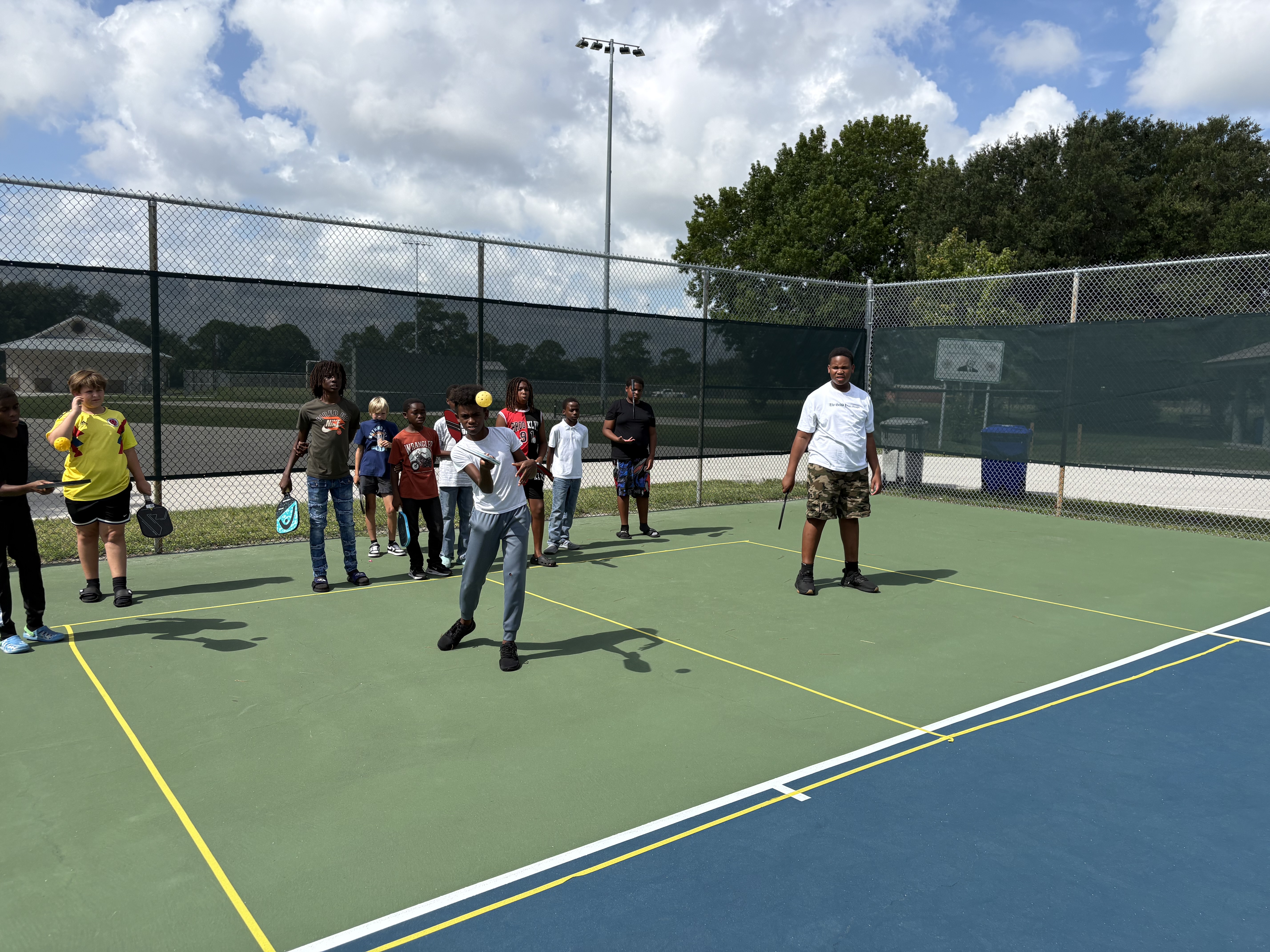 A camper driving a forehand while classmates and a coach watch from the sideline.
