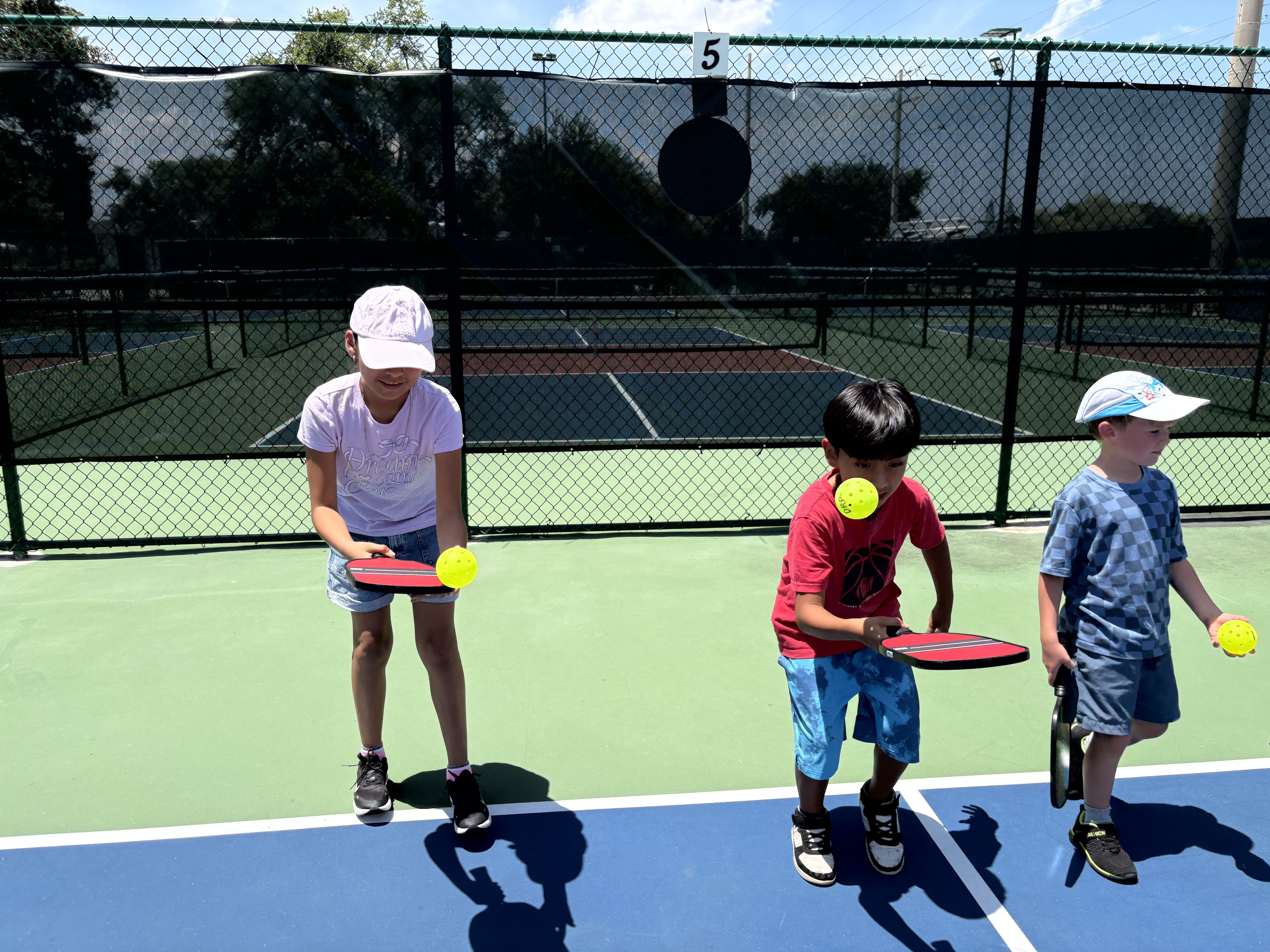 Three young campers practicing balancing balls on their paddles along the baseline.