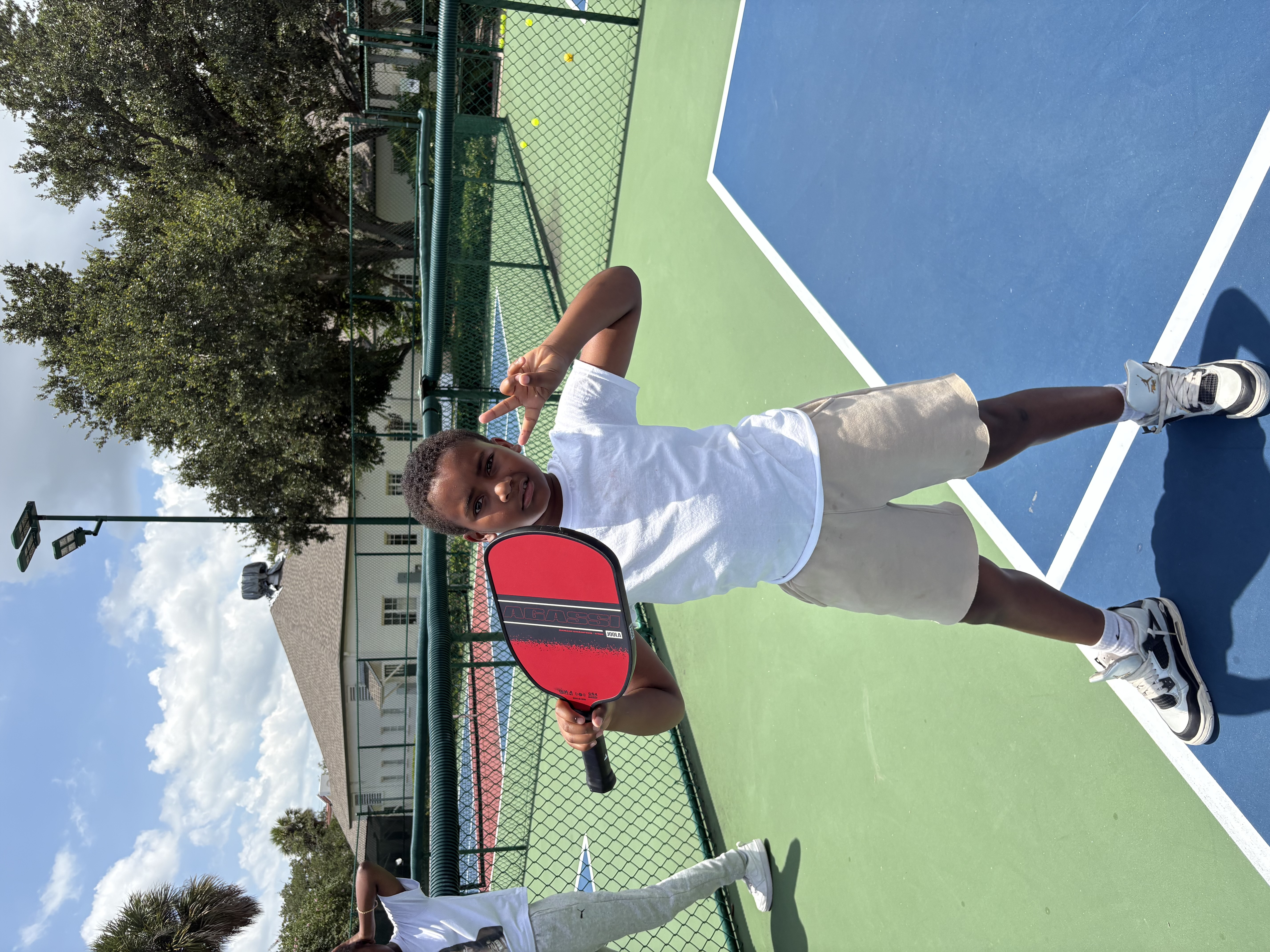 A young player resting a paddle on his shoulder and flashing a peace sign on a sunlit court.