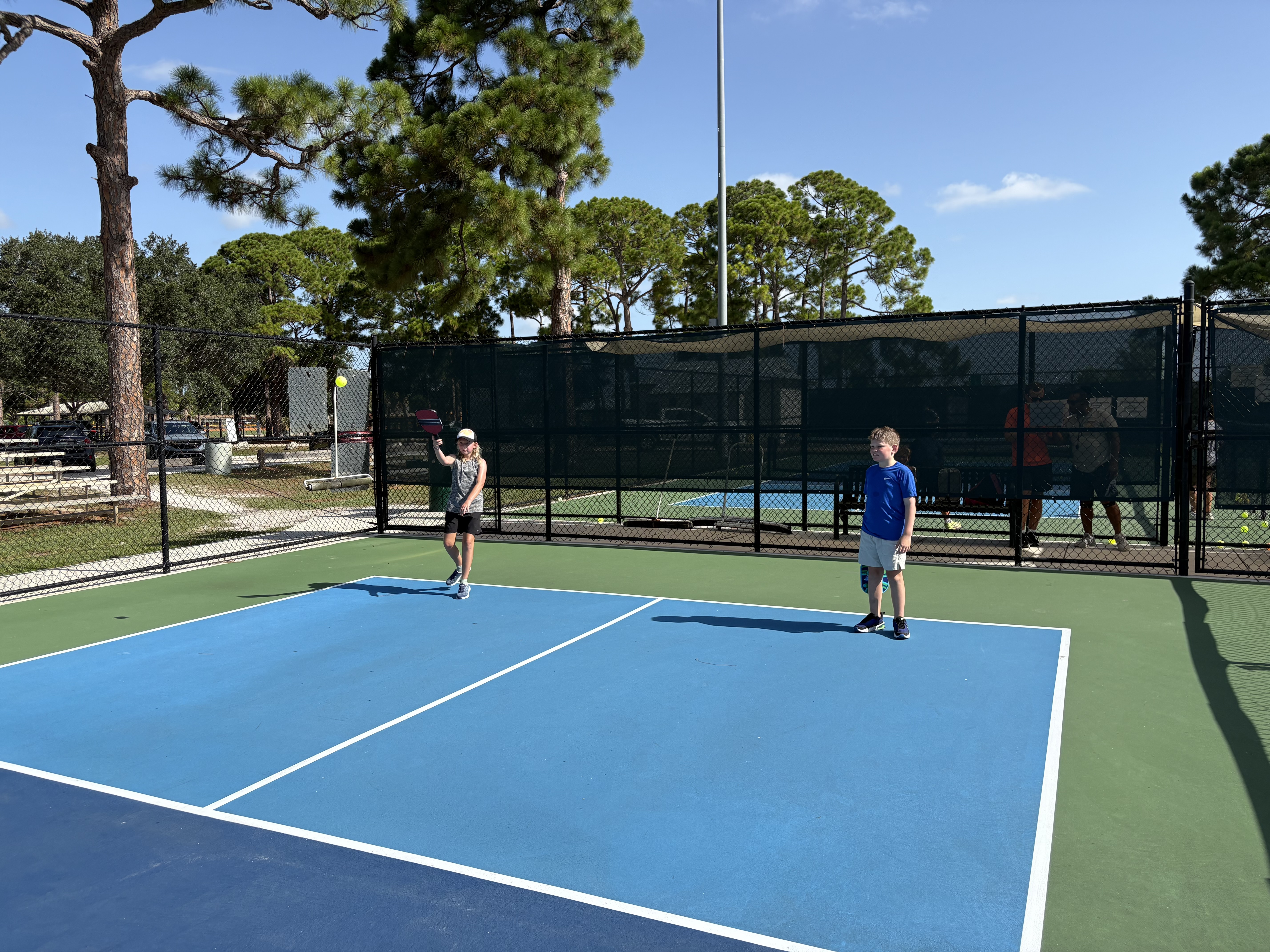 Two campers warming up on an empty court framed by tall pines.